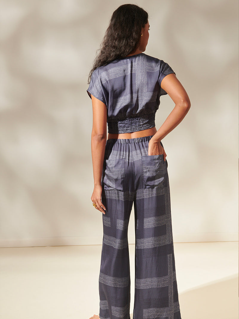 Back view of a woman standing, wearing an Alia Plunge Crop Top and Desta Wide Leg Pants featuring white and charcoal stripes set against a cool steel blue background.