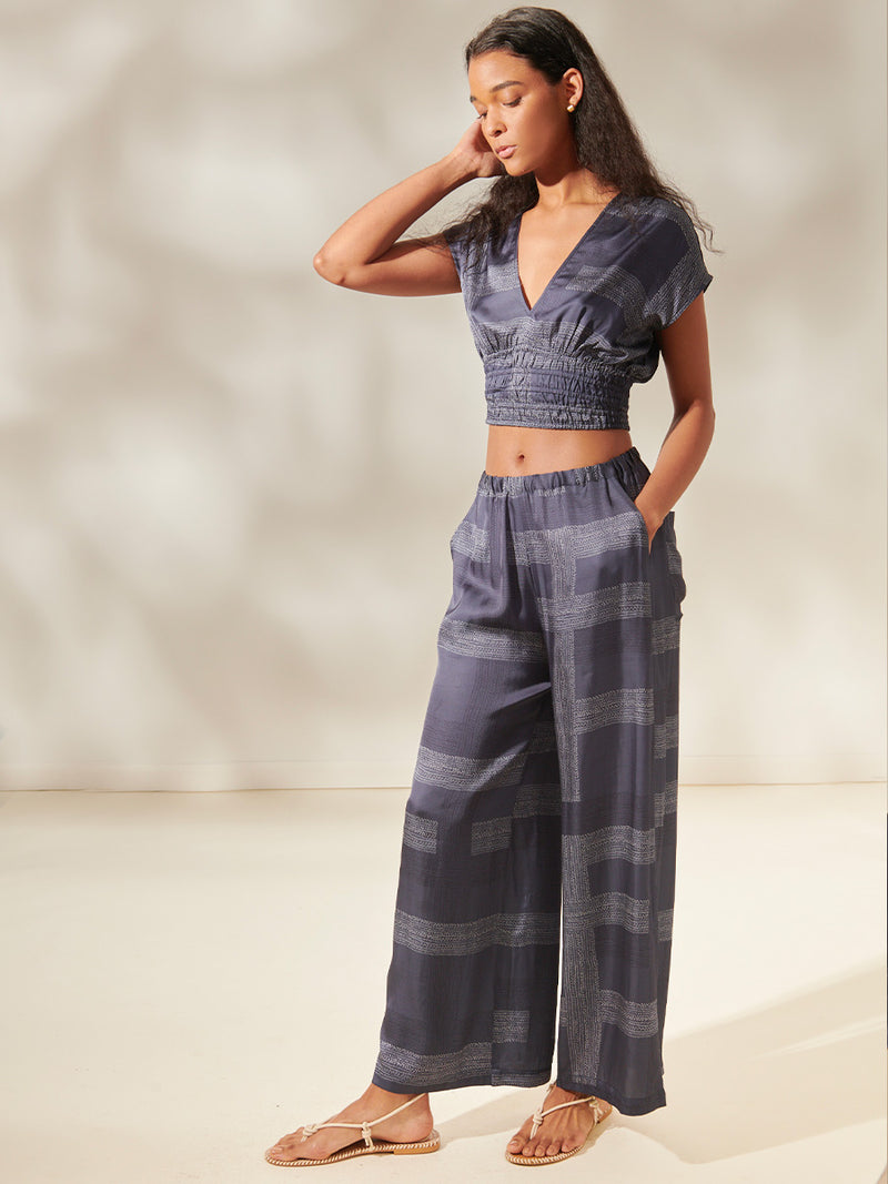 Side view of a woman standing, wearing an Alia Plunge Crop Top and Desta Wide Leg Pants featuring an elegant hand-sketched chevron pattern. The pattern showcases alternating neutral and charcoal stripes set against a cool steel blue background.