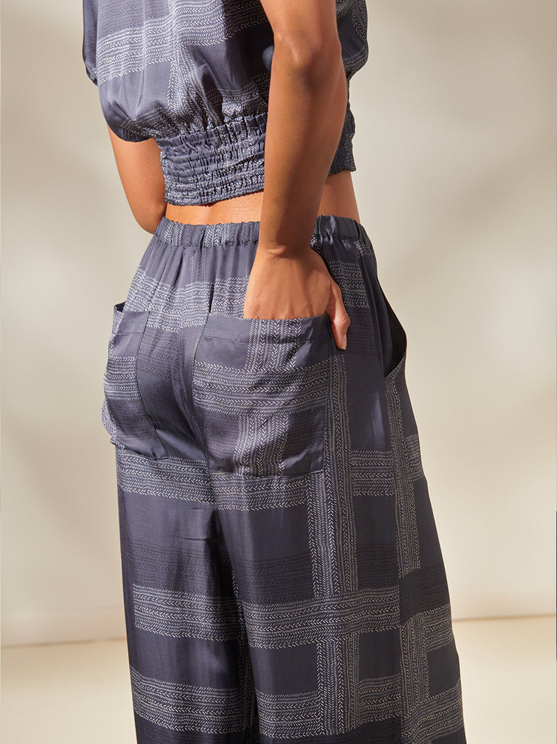 Back view of a woman standing, wearing an Alia Plunge Crop Top and Desta Wide Leg Pants featuring an elegant hand-sketched chevron pattern. The pattern showcases alternating neutral and charcoal stripes set against a cool steel blue background.