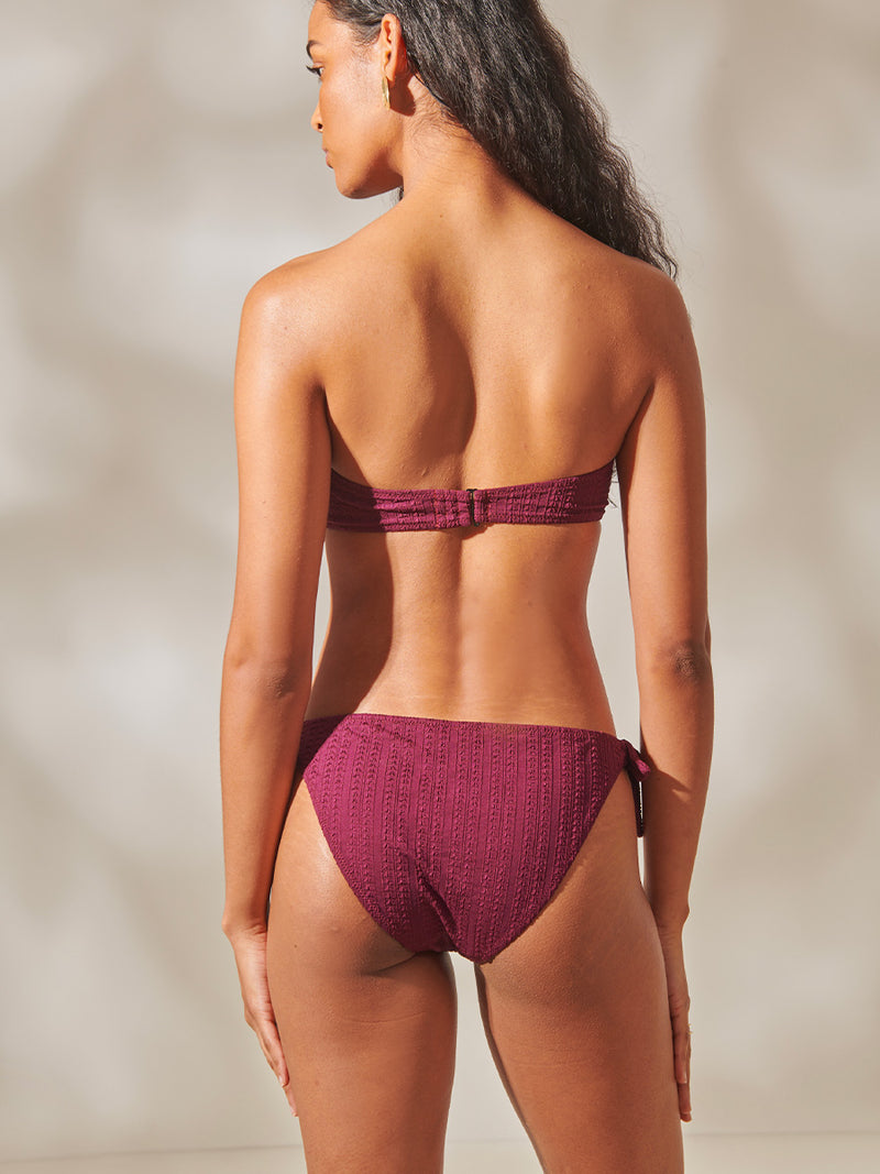 Back of a Woman Standing Wearing Ava Bandeau Top and side tie bikini bottom featuring a downsampled Jordanos Pattern in a luxurious burgundy hue