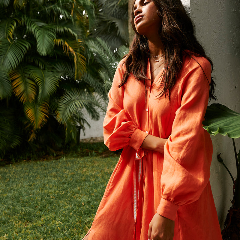  Woman standing with her arm across her her body wearing the lemlem makeda long dress in coral linen tencel. 