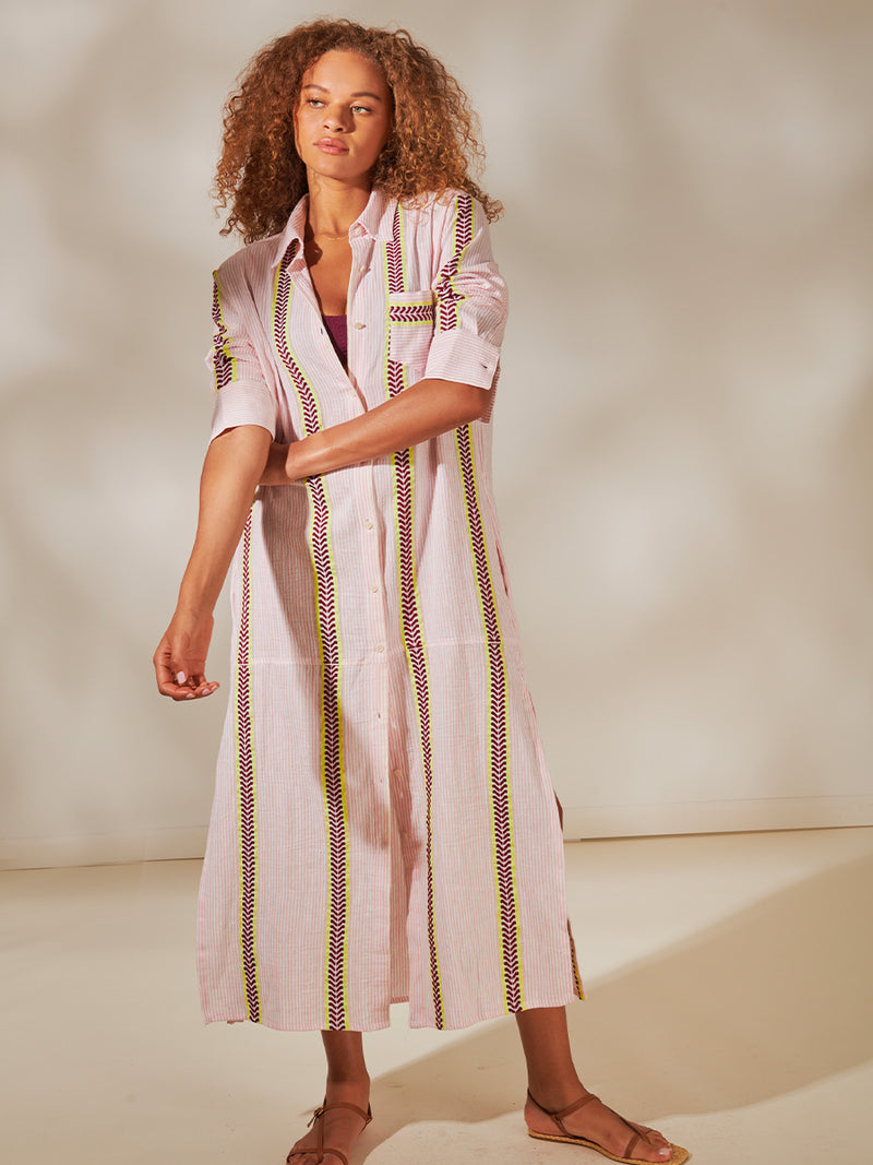 Woman Standing Wearing Anata Shirt Dress featuring delicate pink stripes with a bold chevron patterned ribbon, along with muted hues of pink, burgundy, and a bright citrus-orange hue.