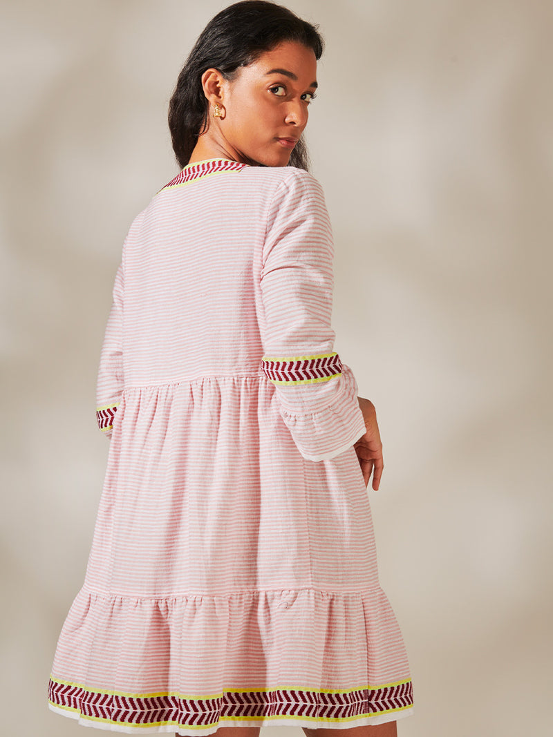 Back View of a Woman Standing Wearing Hanna Flutter Dress featuring delicate pink stripes with a bold chevron patterned ribbon, along with muted hues of pink, burgundy, and a bright citrus-orange hue.