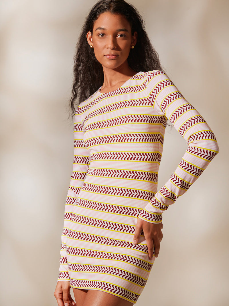 Woman Standing Wearing Nyala Bodycon Dress featuring a range of subtle hues from pink to burgundy with an accent of citron. It features a bold chevron pattern over a delicate pink striped micro terry texture. Contrasting ivory fabric is printed with pink stripes and geometric shapes.