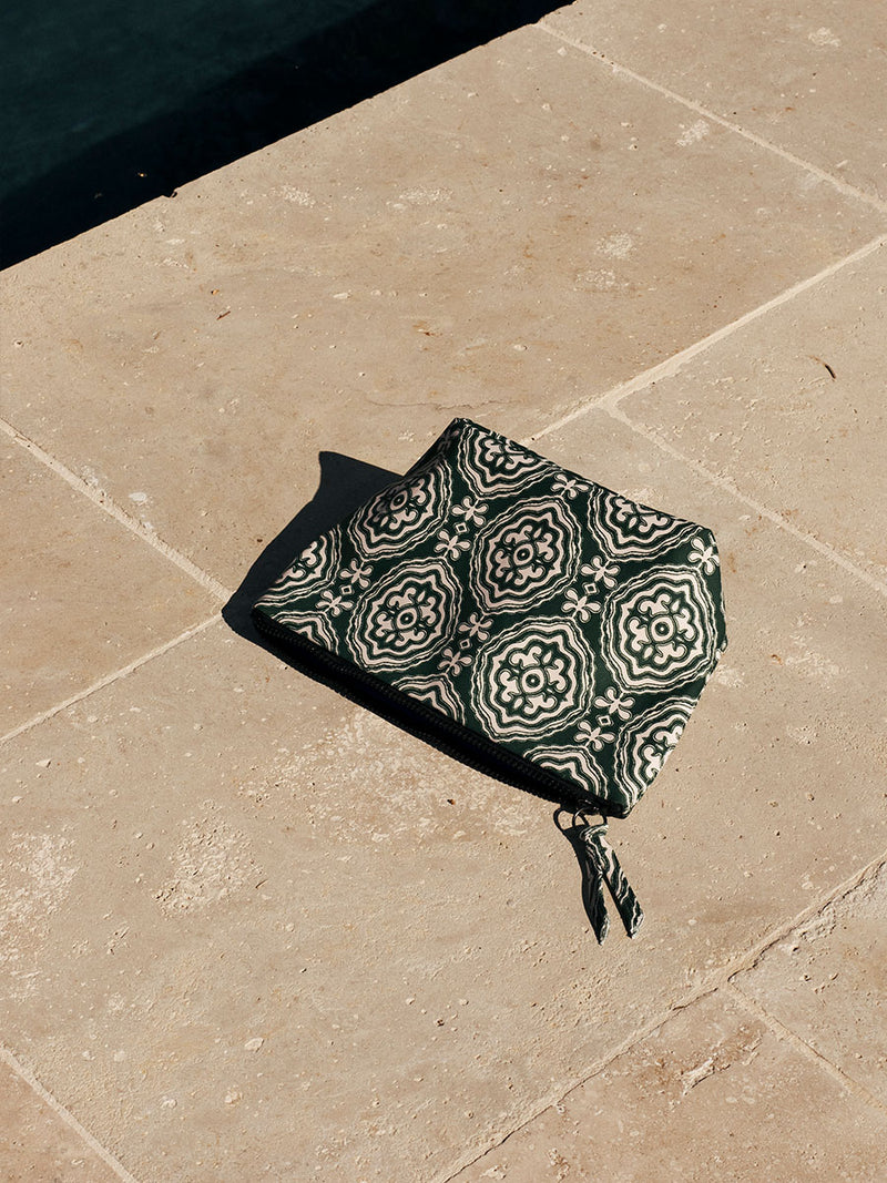 A green travel pouch featuring all over medallion pattern and a black zip closure at the top laid flat on a stone floor by the side of a pool. 