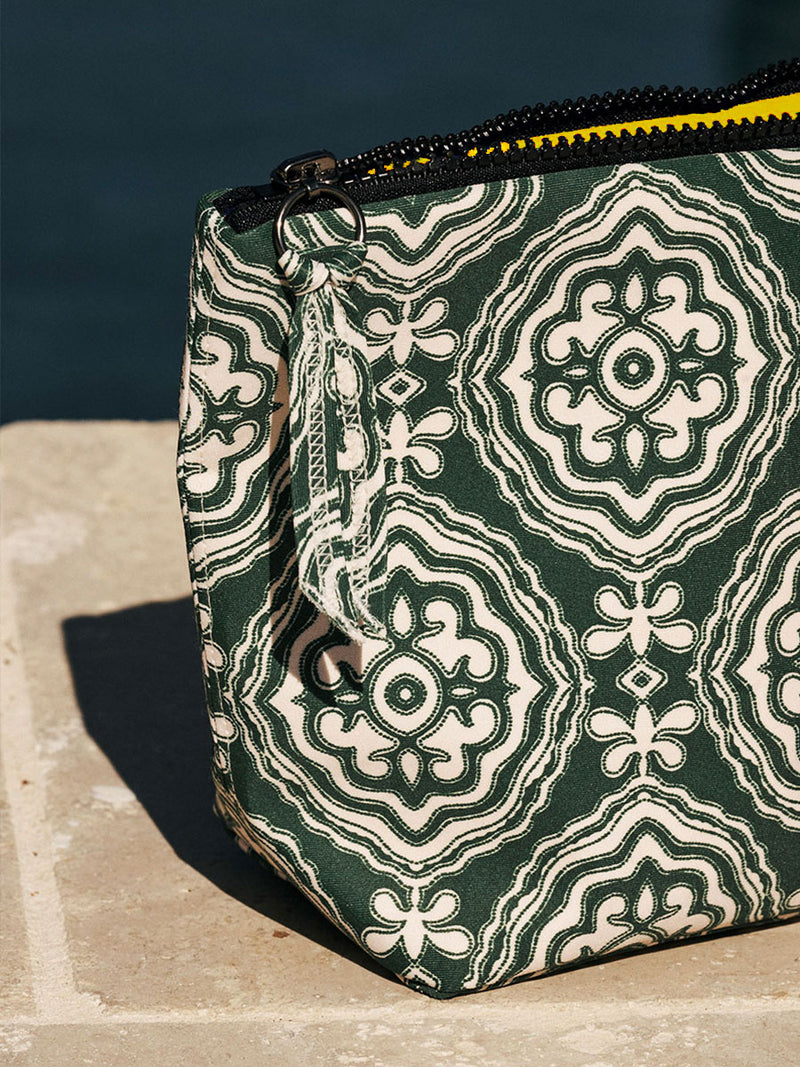 Close up on a green travel pouch featuring all over medallion pattern and a black zip closure at the top on the ground by a pool.