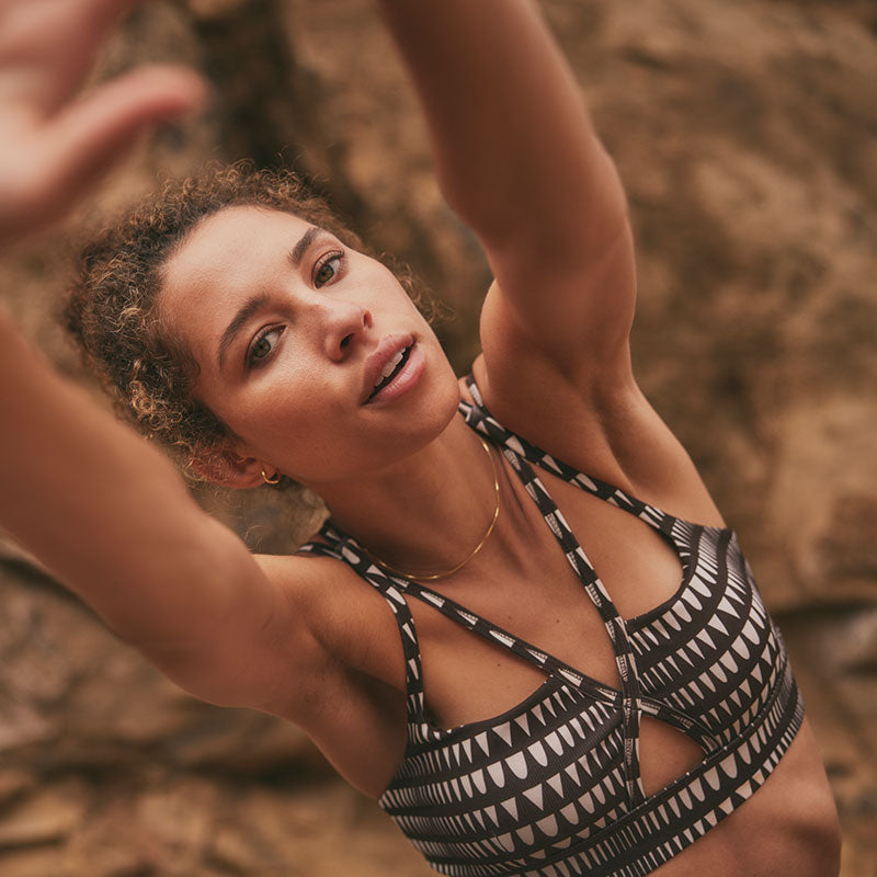 Woman stretching in front of a cliff wearing a Puma x lemlem brown low impact bra.