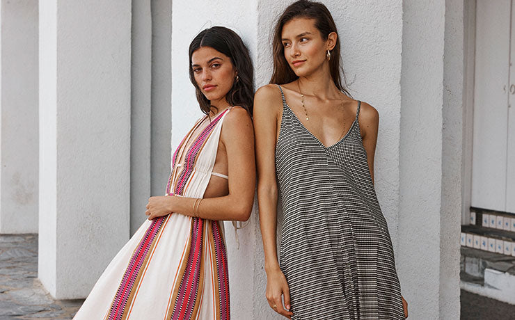 Two women standing against a white wall. One wearing a cut out white with brown and pink stripes lemlem dress. The other one wearing a brown lemlem slip dress. 