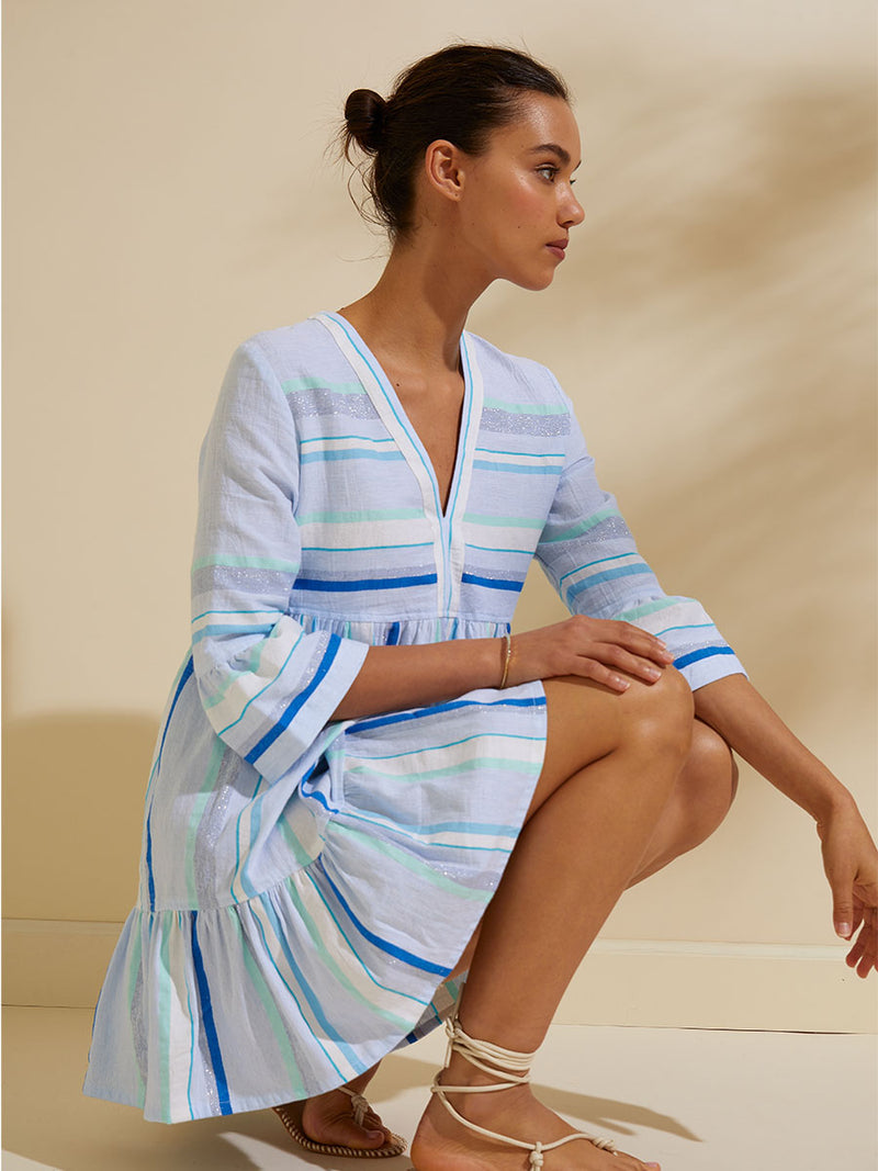 Side view of a woman squatting wearing the Ruki Flutter Dress featuring a mutli tonal stripe pattern in five shades of blue with silver and white accents.