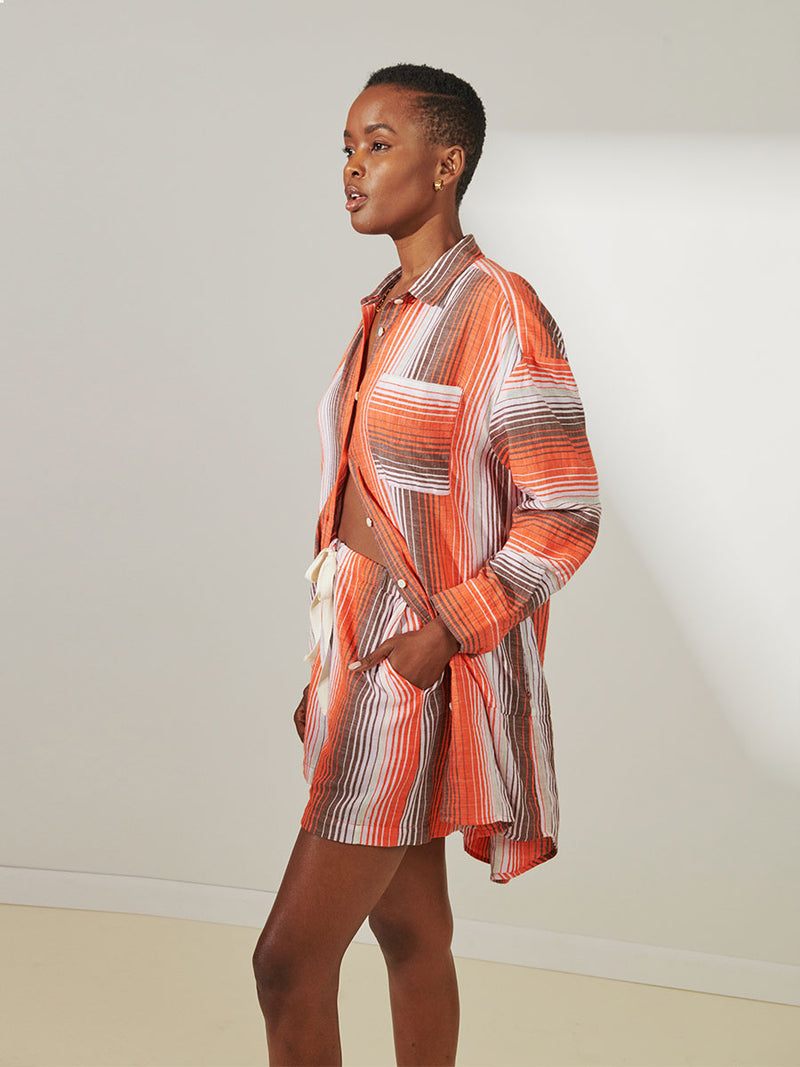 Side View of a Woman Standing Wearing lemlem Mariam Shirt featuring graded continuous stripe pattern creating an ombre effect featuring earth, orchid & burnt orange and matching shorts