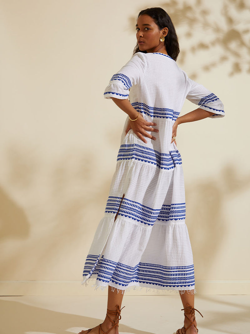 Back view of a woman standing with her hands on her waist wearing the Yani Long Flutter Dress featuring blue tibeb diamond design bands on a textured seersucker white background.  