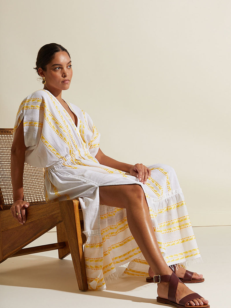 Woman seating on a chair wearing the Abeba Plunge Neck Dress featuring the yellow signature Tibeb pattern and gold lurex highlights on a white background.