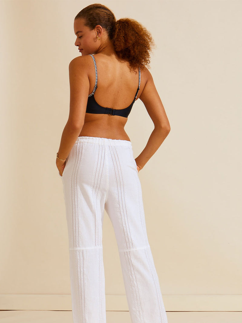 Back view of a woman standing with her hand in her pocket wearing the Abira Drawsting Pants in white with silver lurex, worn with the black Lena bralette swim top