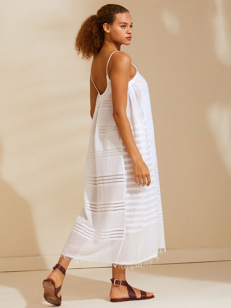 Side view of a woman walking wearing a white Abira long slip dress with spaghetti straps and stitches of silver lurex