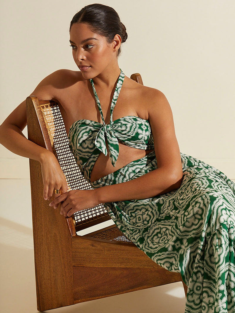 Woman sitting on a chair wearing the Medallion Cutout Dress featuring architectural white patterns on a deep green background.
