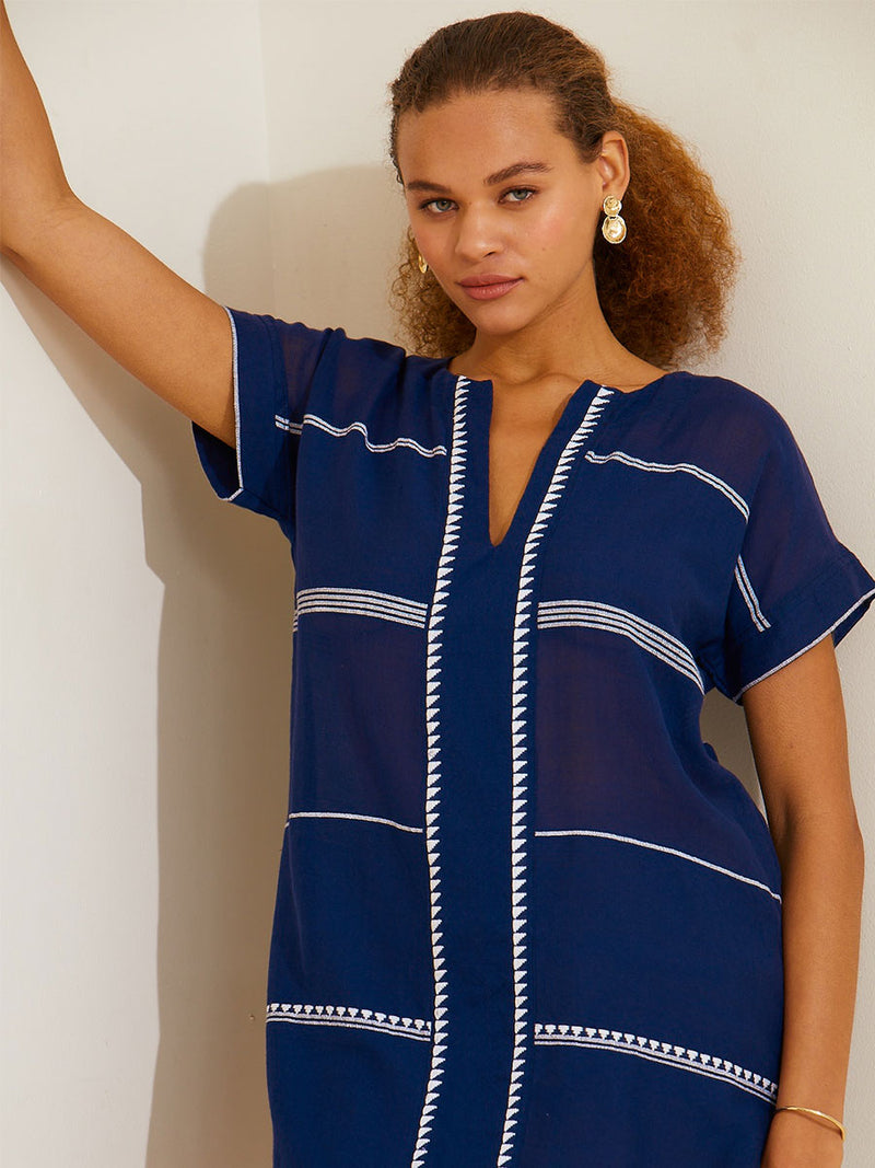 Woman standing with her arm up on a white wall wearing the Nunu Tunic Dress in navy blue featuring white stripes and graphic lines
