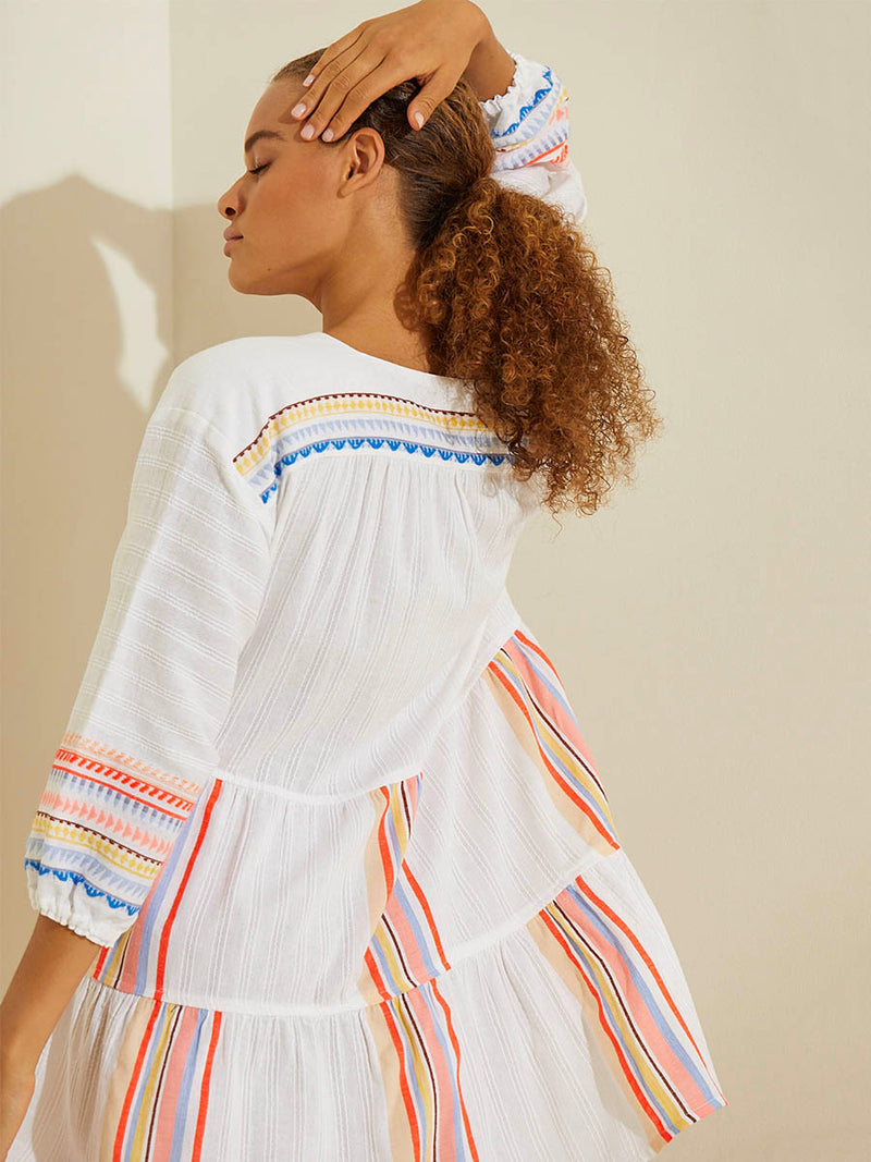 Back view of a woman standing with her hand on her head  wearing the Bekah Popover Dress featuring 10 tutti frutti colors embroidered on a white background.  