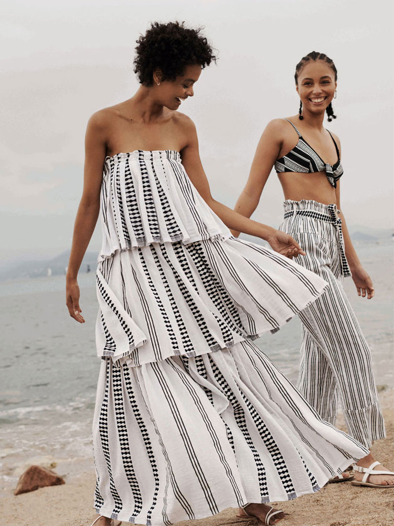 Two woman dancing on a beach one wearing a white lemlem off the shoulder dress with graphic black lines and the other wearing the Luchia Front tie Bikini top with matching white pants with black graphic lines. 