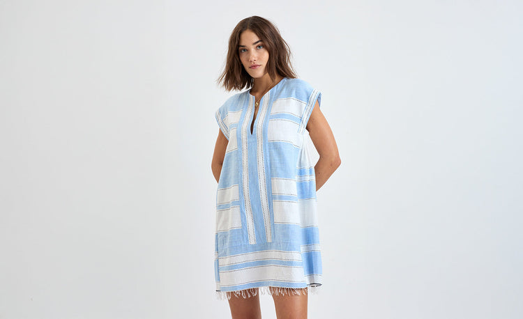 Woman standing with her hand by her neck wearing a handwoven lemlem short plunge neck dress in light peach and pink gauze.