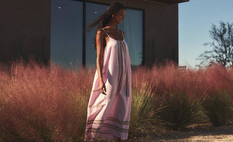 Woman Sitting on the beach wearing lemlem handwoven dress featuring  striped pattern in multi colors on cream bav