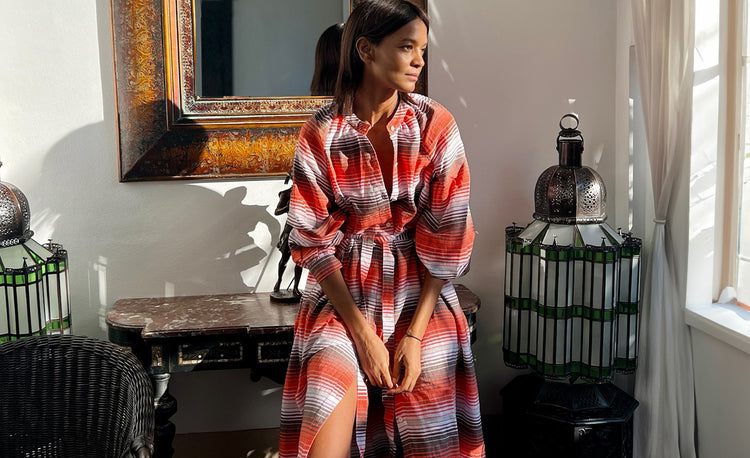 Liya Kebede sitting in an appartment by a window wearing a long handwoven lemlem dress with white, red and brown thin vertical stripes.