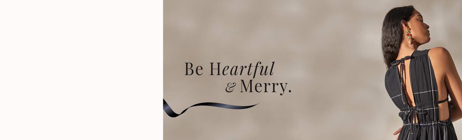 Holiday Message "Be Heartfelt & Merry" on a beige background underlined with a black ribbon and with a woman standing back facing wearing a black dress.