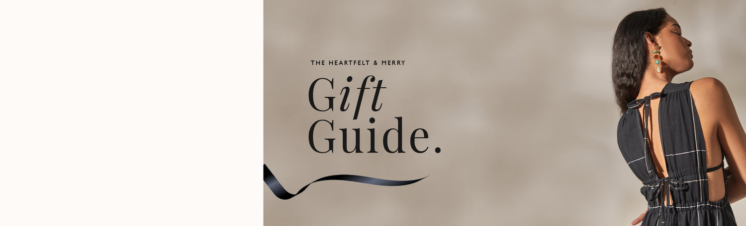 Holiday Message "Be Heartfelt & Merry Gift Guide" on a beige background underlined with a black ribbon and with a woman standing back facing wearing a black dress.