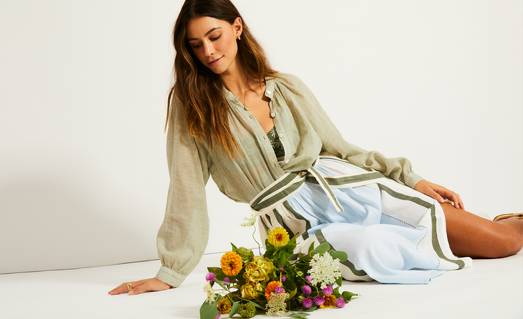 Woman sitting on the flower with a bouquet of flowers next to her. she is wearing a lemlem handwoven blouse in sage with a lemlem handwoven skirt in white with green and blue stripes.