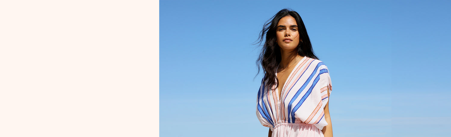 Woman standing against a blue sky and wearing an pale pink Eskedar plunge neck dress with blue and coral stripes