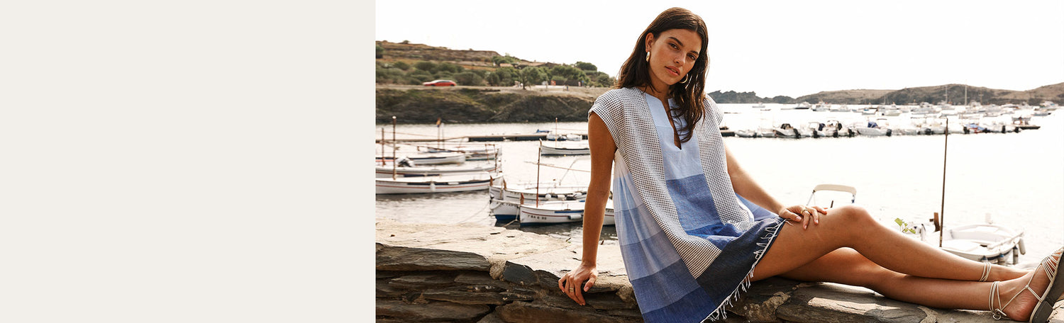 Woman lounging on a stone wall with a sea port in the background, wearing a short cafta dress in blue and and white patchwork.