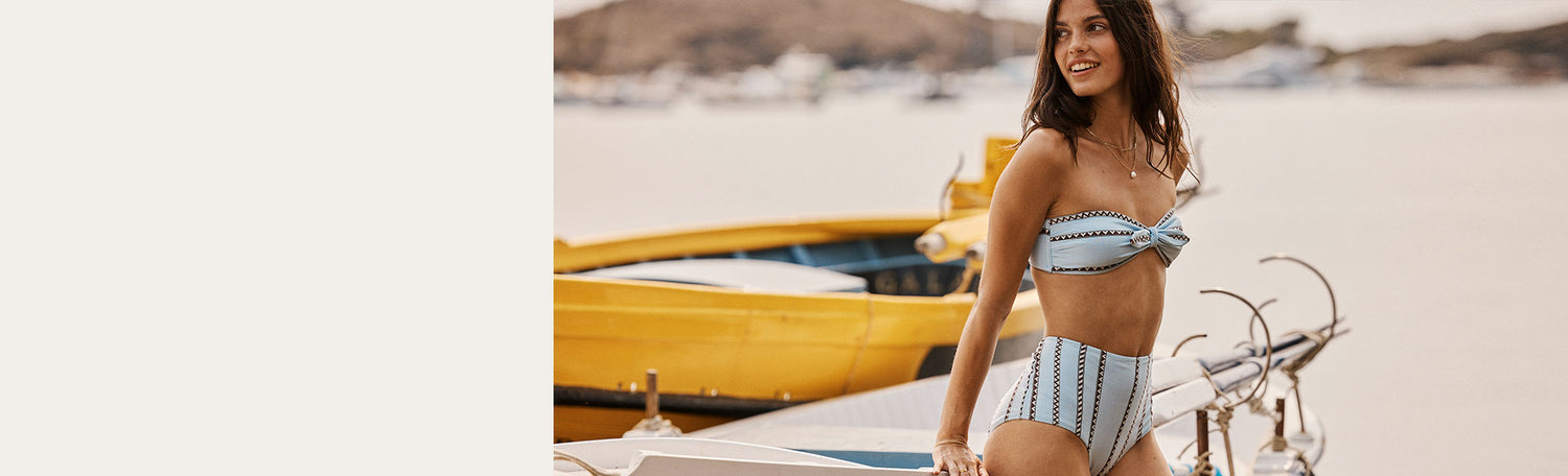 Woman leaning against a small fishing boat wearing a blue with brown graphic strips bandeau bikini top and matching high waist bottom.