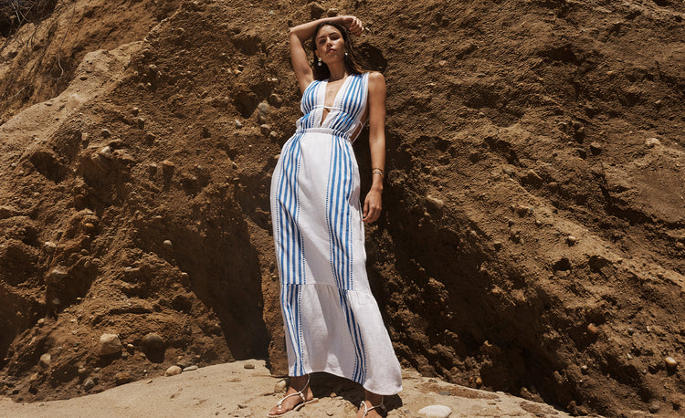 Woman standing on a beach in front of a cliff wearing a long lemlem hand woven dress in white with vertical blue stripes.