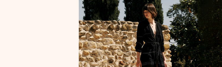 Woman standing in front an old stone wall and trees wearing a black long sleeve robe