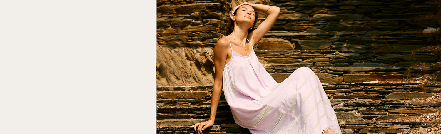 Woman sitting a rocky stairs pulling her hair around her head and wearing a light pink long slip dress.
