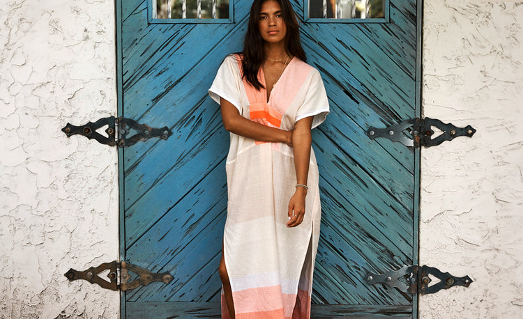 Woman standing against a blue wooden door wearing a lemlem handwoven cafta dress in white with orange color block around the neck and hem