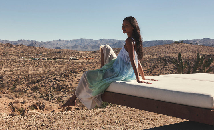 Liya Kebede Walking on the beach wearing lemlem handwoven slip flowy dress featuring ombre pattern in navy, blue, pale blue and white colors