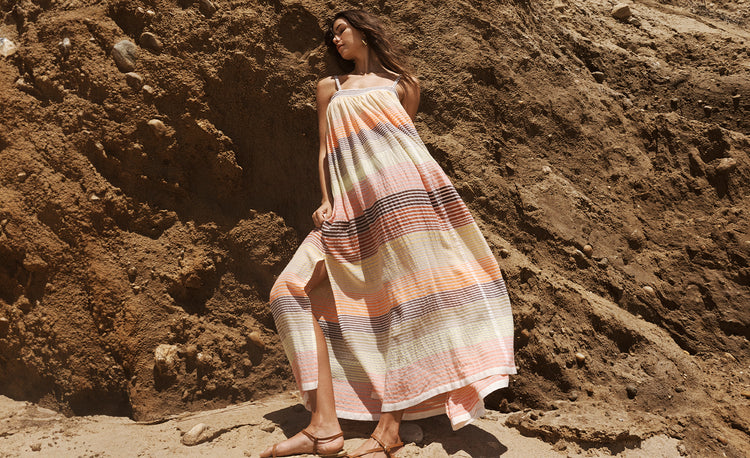 Woman Standing wearing lemlem Amaresh Sunrise Slip Dress featuring stripe pattern in brown, orange, yellow and white colors