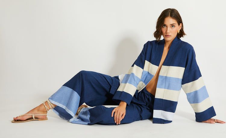 Woman Sitting wearing lemlem Jacket featuring color block pattern in blue. cream and white colors and pants in dark blue color with pale blue accents