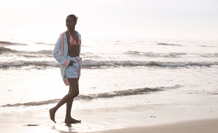 Woman Walking on the beach wearing lemlem Airi Sky Shirt and matching shorts featuring sky blue color with neon orange pops and triangle bikini top 