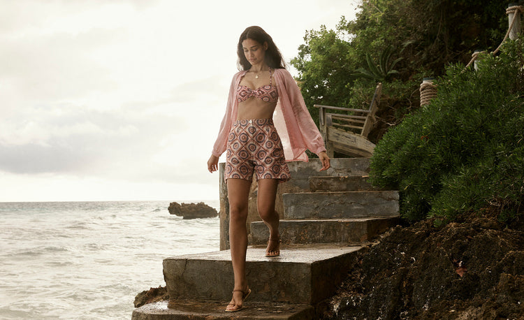Woman Walking on the beach wearing lemlem Bikinis and Shorts featuring pattern, blending peach, rust, and orchid hues with a bold citrus yellow pop and sheer blouse in rose color