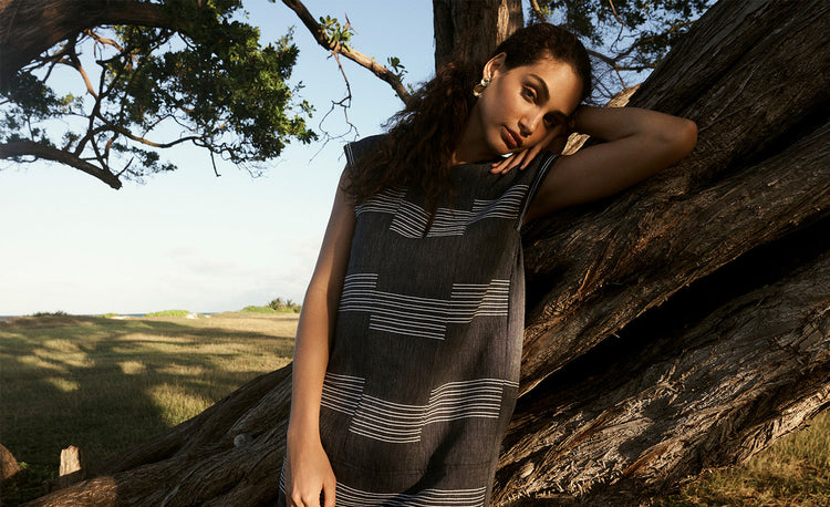 Woman Standing Leaning on the tree in Jamaica wearing lemlem Caftan dress featuring navy color and stripe pattern in white color