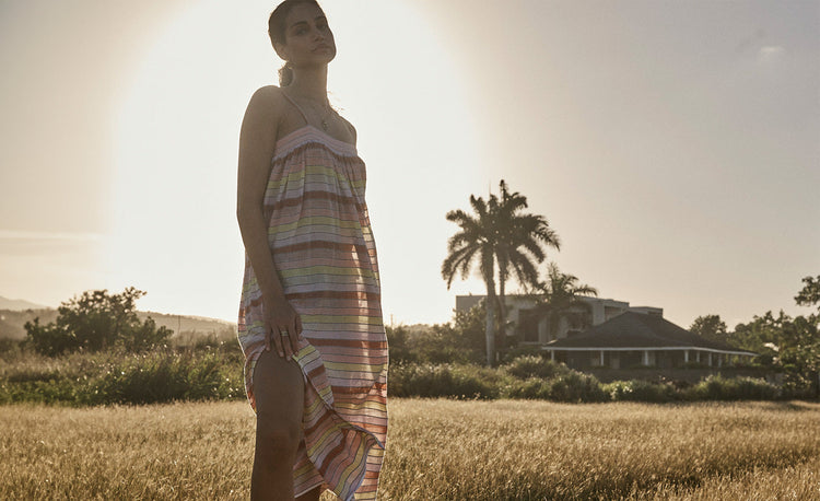 Woman Standing with the sunset on the background, wearing lemlem Flowy Dress featuring featuring soft sorbet-colored stripes in peach, rust, and orchid, accented with a vibrant citrus yellow pop