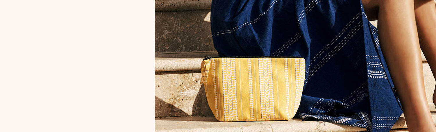 Yellow beach pouch on stone stairs standing next to a woman's legs. The woman is wearing a navy blue handwoven skirt.