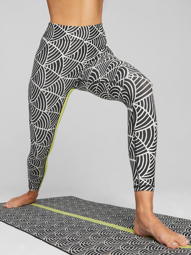 Woman Exercising on a yoga mat wearing Puma x lemlem Leggings in Ghost Pepper and Black colors  and matching Puma x lemlem low impact bra  featuring hand sketched scallop print and color block accents in bright pink and green colors.