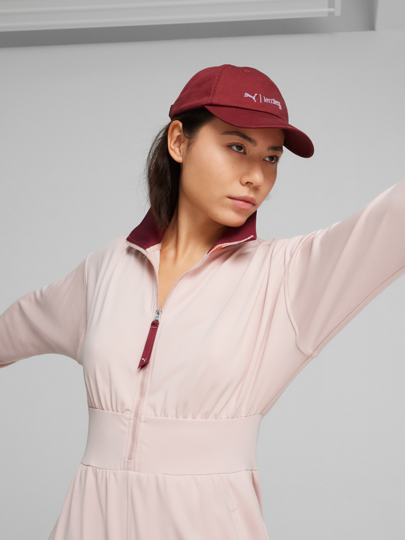 Woman Standing Wearing Puma x lemlem Twill Cap in Regal Red Color and a Jumpsuit in rose quartz color