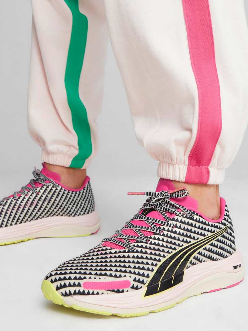 Close up on a legs of a woman standing wearing Puma x lemlem Velocity Nitro Sneakers featuring tibeb tirangle print., frosty rose and frosty pink colors with black and yellow color block details.