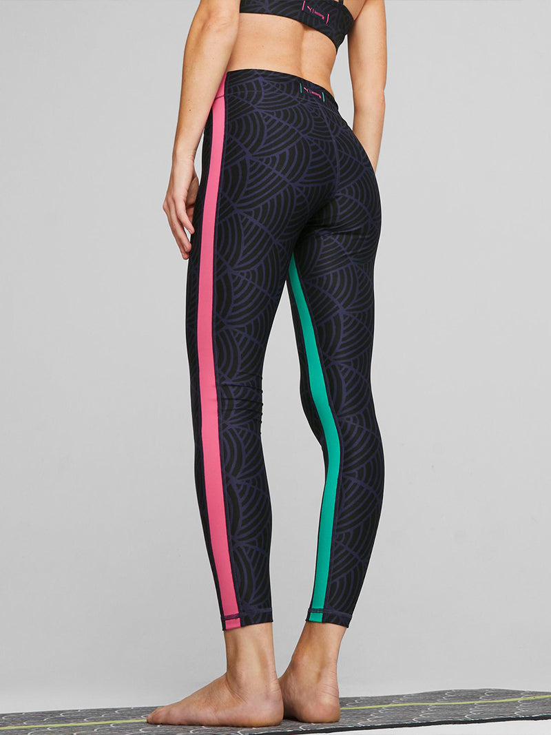 Woman Standing  on a yoga mat wearing Puma x lemlem Leggings in Navy and Black colors  and matching Puma x lemlem low impact bra  featuring hand sketched scallop print and color block accents in bright pink and turquoise colors.