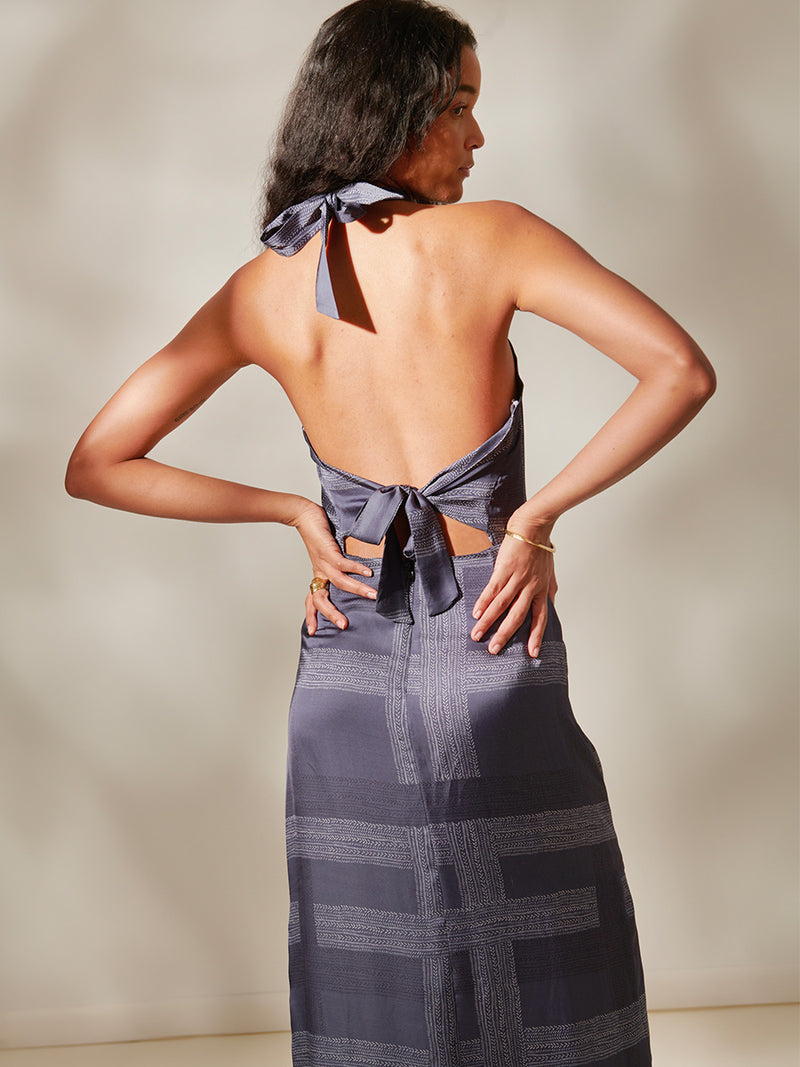 Back of a Woman Standing Wearing Ajani Halter Dress Featuring an elegant hand-sketched chevron pattern, featuring alternating neutral and charcoal stripes set against a cool steel blue background.