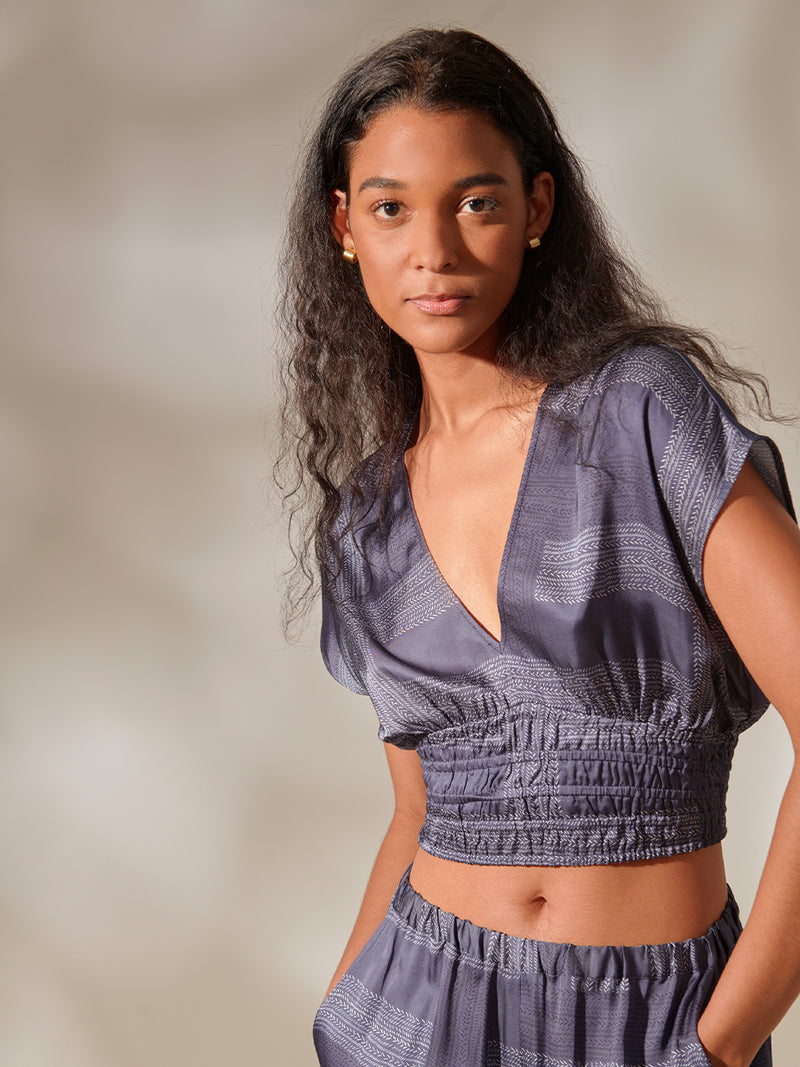 Close up of a Woman Standing Wearing Alia Plunge Crop Top and Desta Wide Leg Pants featuring white and charcoal stripes set against a cool steel blue background.
