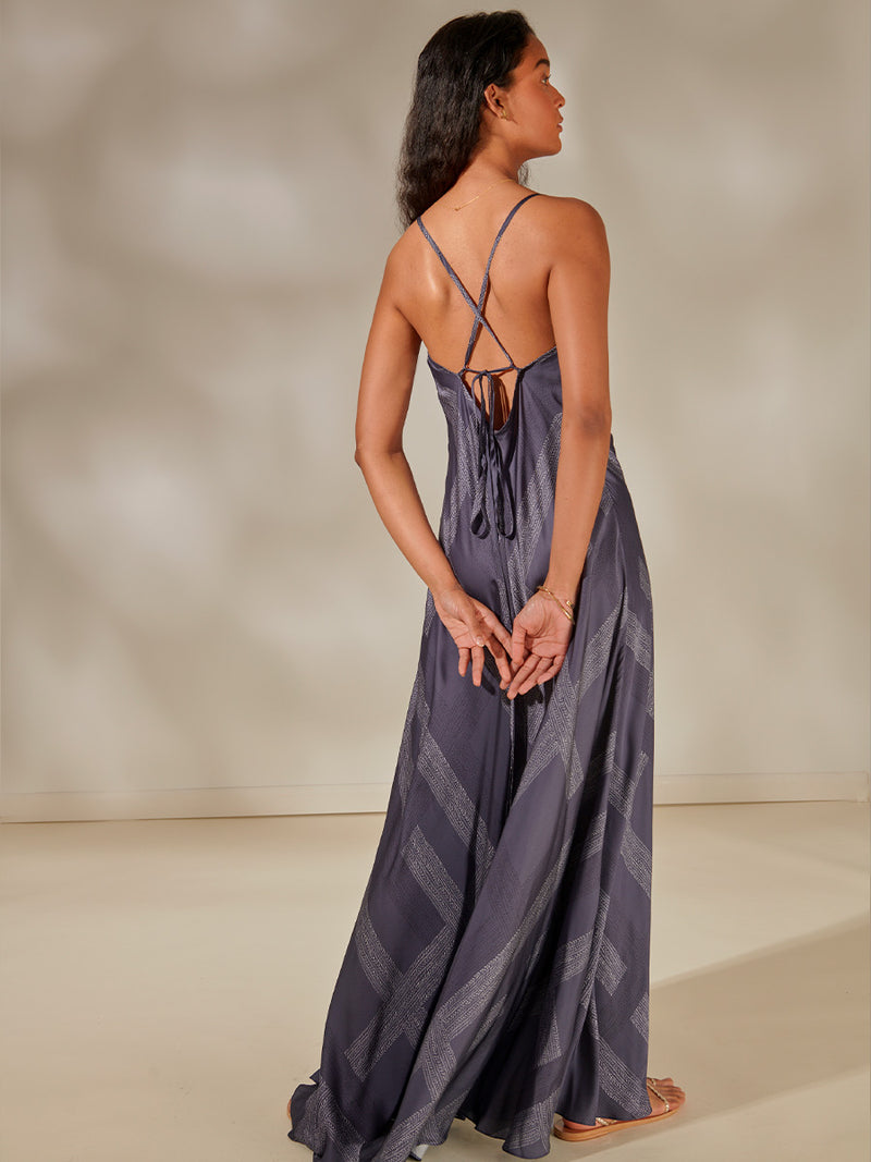 Back of a Woman Standing Wearing Aluna Slip Dress featuring an elegant hand-sketched chevron pattern, featuring alternating neutral and charcoal stripes set against a cool steel blue background.